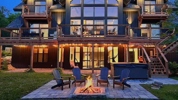 A large, multi-story house with a deck, outdoor seating, and a fire pit. - Laurentian Chalet: 4K Cinema, Sauna & Hot Tub (Morin-Heights)