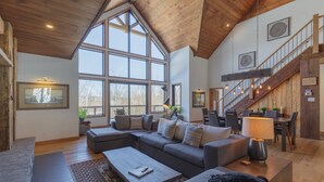 A spacious living room with a large window, a staircase, and a cozy seating area. - Laurentian Chalet: 4K Cinema, Sauna & Hot Tub (Morin-Heights)