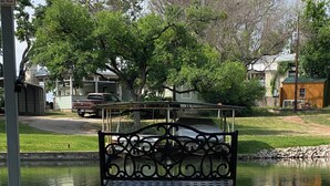 A bench with intricate metalwork, positioned in front of a calm water body, likely a pond or small lake, with a residential area in the background. - Hidden Harbor-Lakefront on Secluded Cove w/Dog Run (Kingsland)