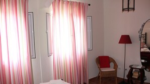 A bedroom with striped curtains, a wicker chair, a red lamp, and a bedside table with books. - Cozy House (Lagos)