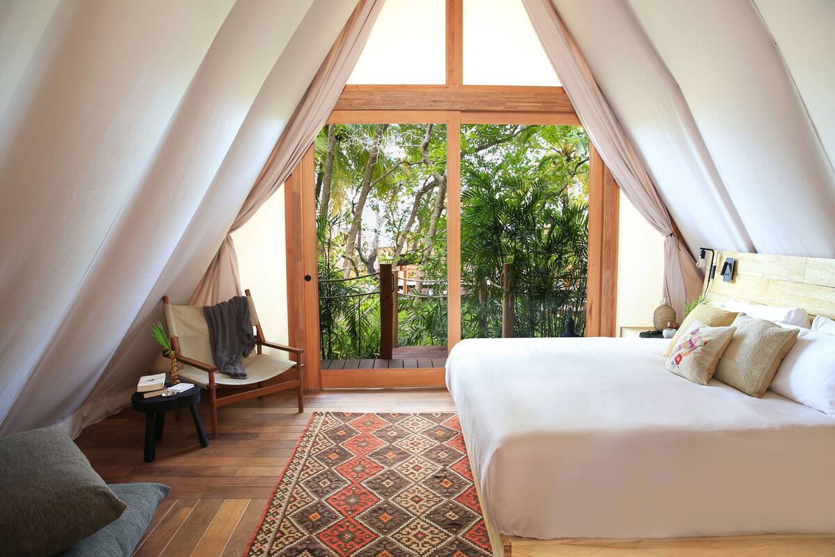 A cozy bedroom with a large bed, a chair, a small table, and a view of lush greenery through a sliding door.