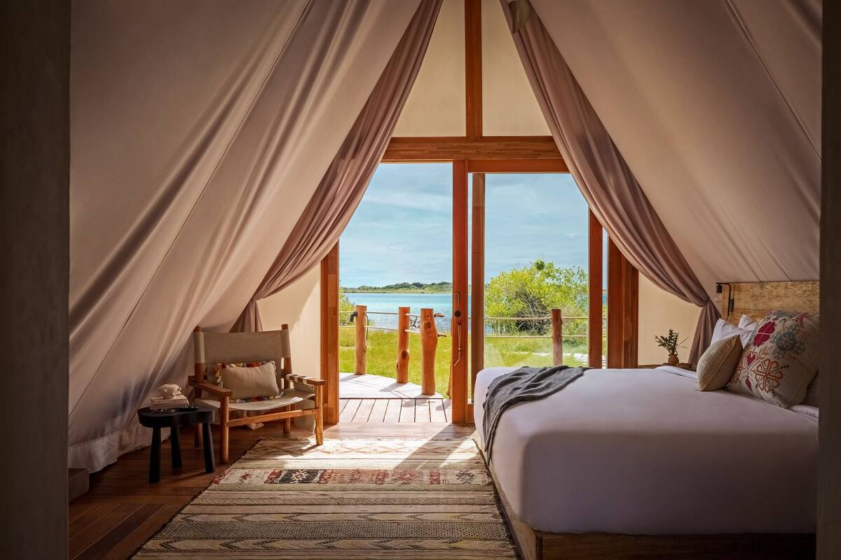 A bedroom with a large bed, a wooden chair, a small table, and a view of the outdoors.
