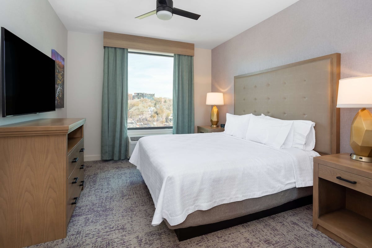 Ein Hotelzimmer mit einem Bett, einem Fernseher, einem Schreibtisch, einer Lampe und einem Fenster mit Vorhängen.
