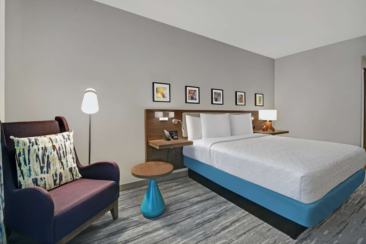 Habitación de hotel con cama, una silla, una mesita, una lámpara y una mesita de noche con teléfono y otra lámpara.