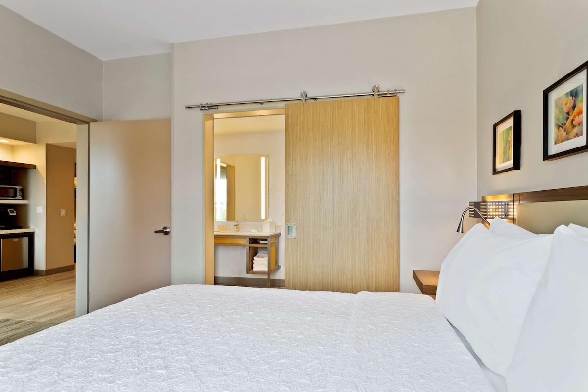 Una habitación de hotel con una cama, una puerta de madera, un baño con lavamanos y una mesita de noche con lámpara.