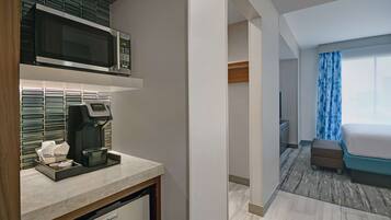 A hotel room with a kitchenette, a bed, a window with curtains, and a view of the city.