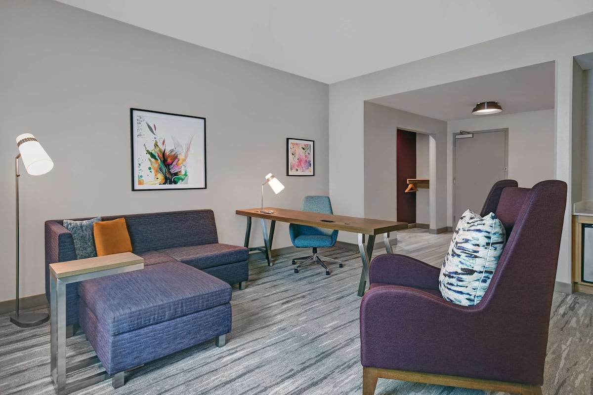 Una habitación de hotel moderna con sofá, sillón, reposapiés y una mesita. Hay dos cuadros enmarcados en la pared.