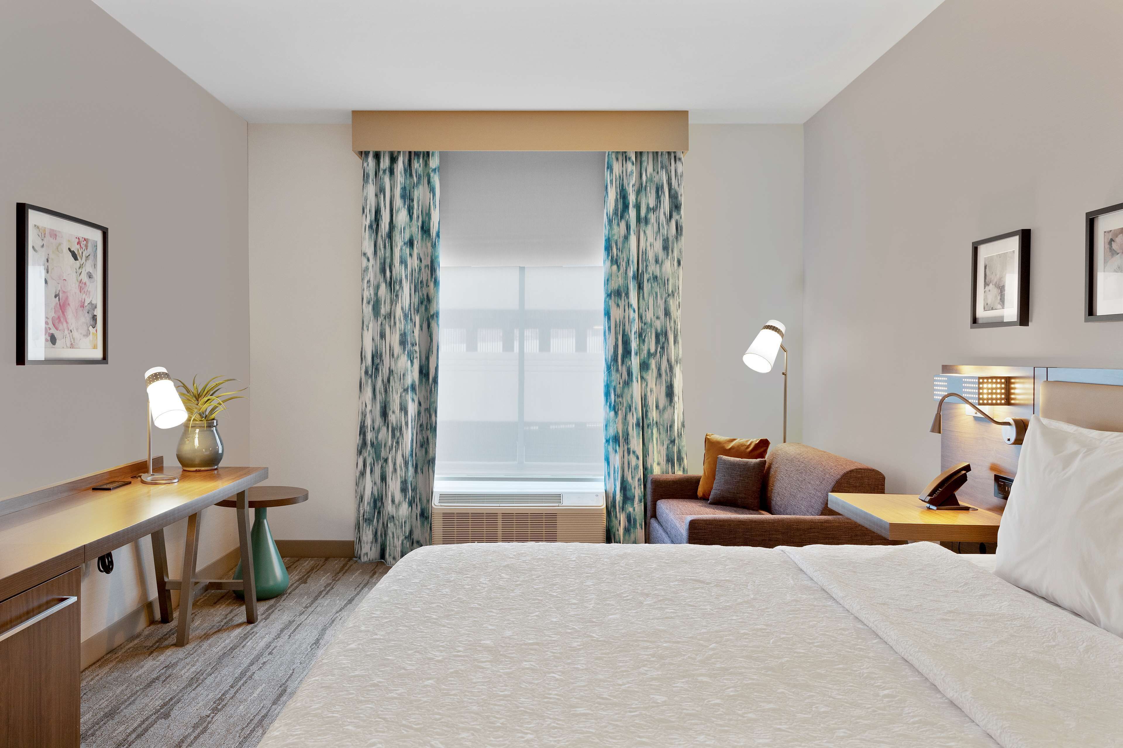 A hotel room with a bed, a desk, a chair, a nightstand, and a window with curtains.