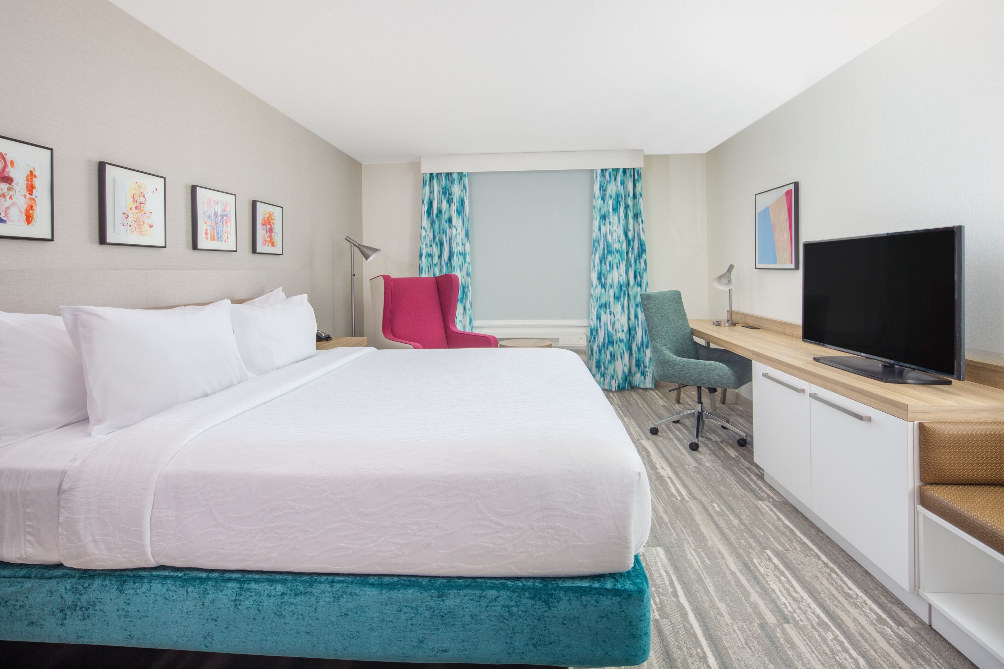 A hotel room with a large bed, a desk with a flat-screen TV, a red chair, and artwork on the walls.
