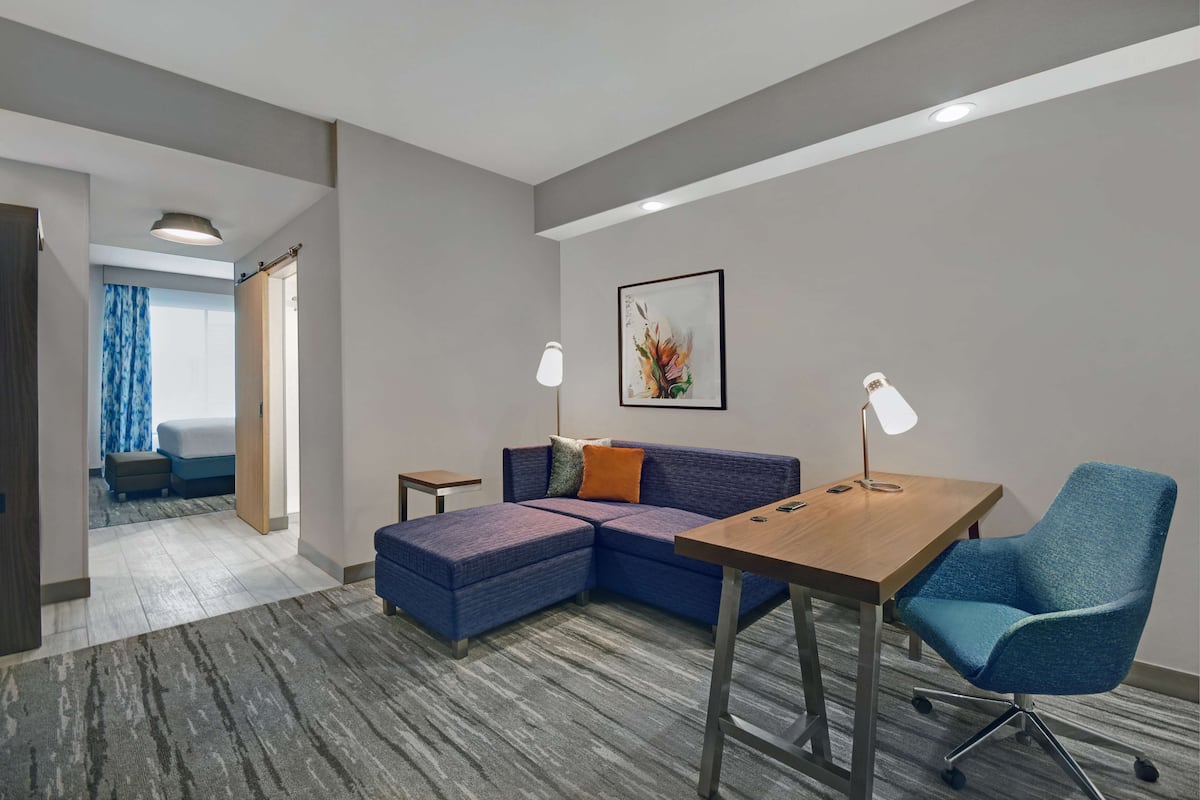 Una habitación de hotel moderna con un sofá modular, un escritorio de madera y un sillón azul. Se observa una obra de arte enmarcada en la pared y una pequeña mesita.