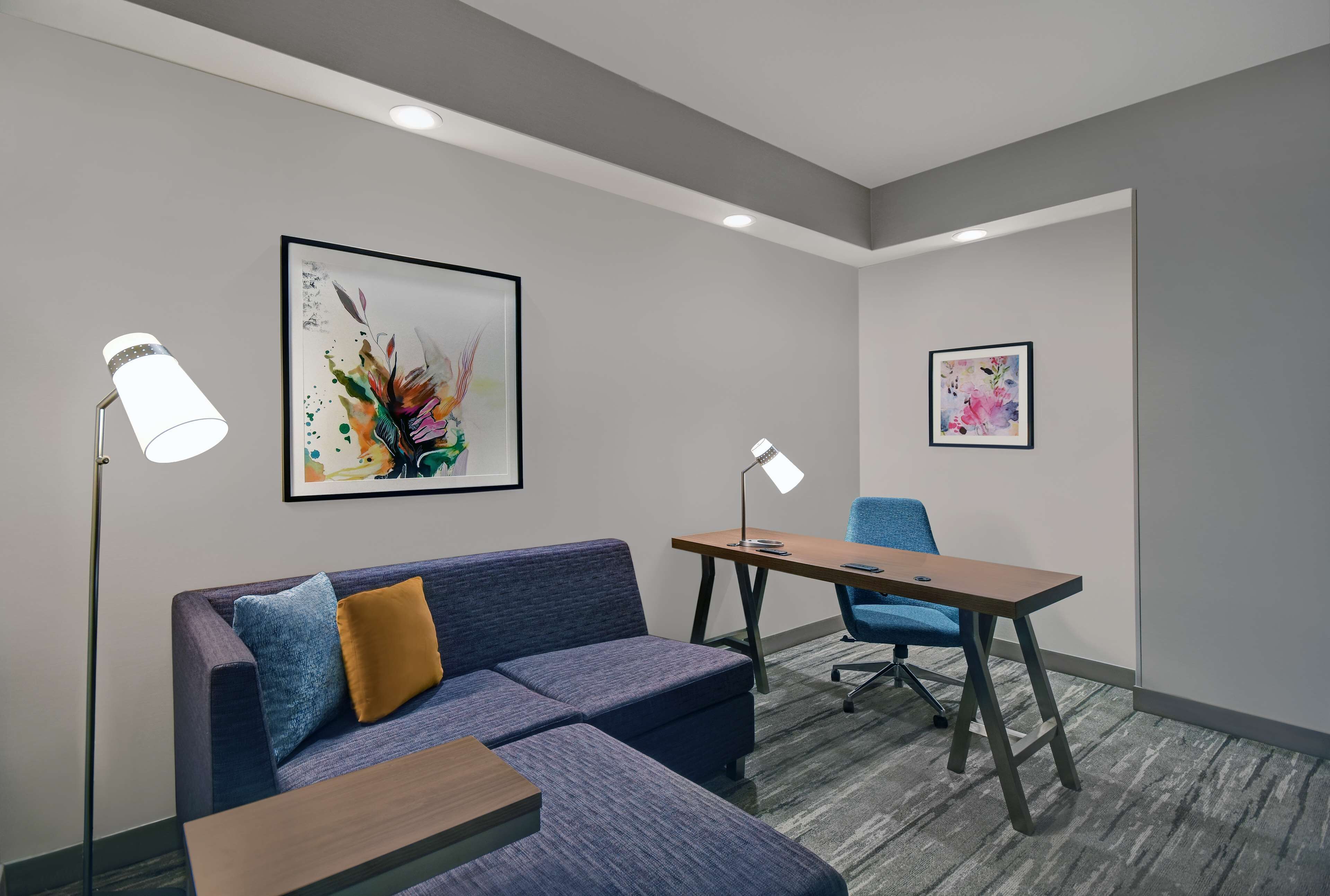 A modern meeting room with a blue sofa, a wooden table, and two chairs. There are two framed artworks on the walls and two floor lamps.