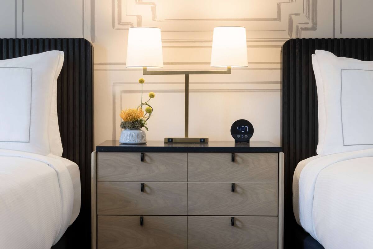 Een hotelkamer met twee bedden, een nachtkastje met een lamp, een klok en een kleine vaas met bloemen.
