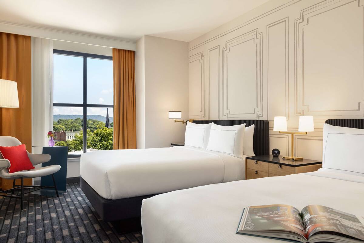 Hotelkamer met twee bedden, een bureau en een stoel. Er is een raam met uitzicht op groen en gebouwen.