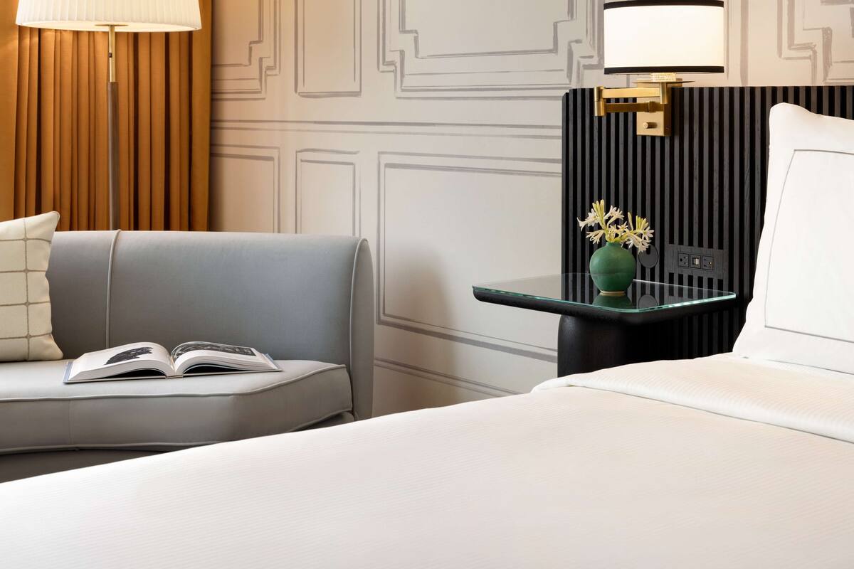 Een hotelkamer met een bed, een slaapbank, een nachtkastje met een lamp en een vaas met bloemen.