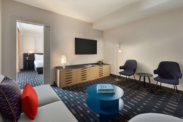 Habitación de hotel con sofá, dos sillones, una mesita, televisor, armario y alfombra estampada.