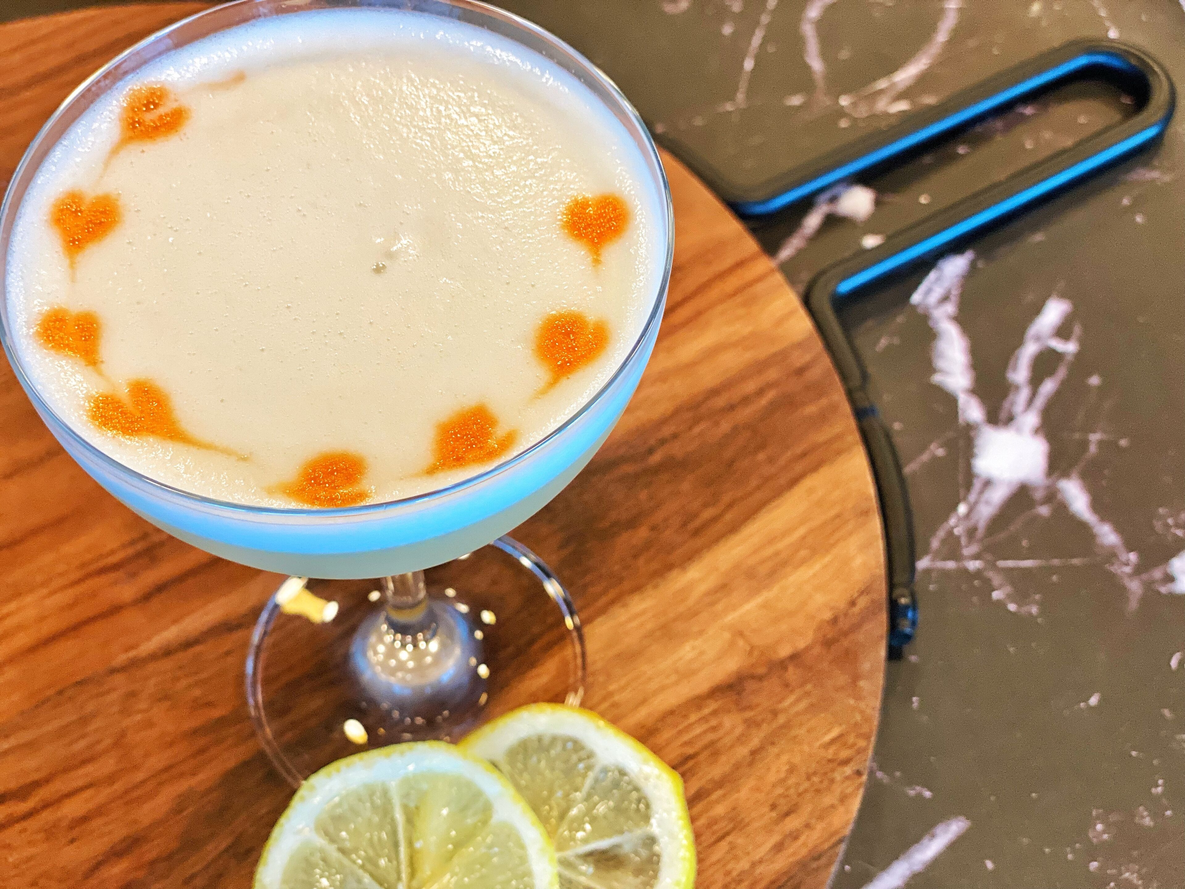 En cocktail med hjerteformede pynt og blå kant, ledsaget af et citronskive på en træoverflade.
