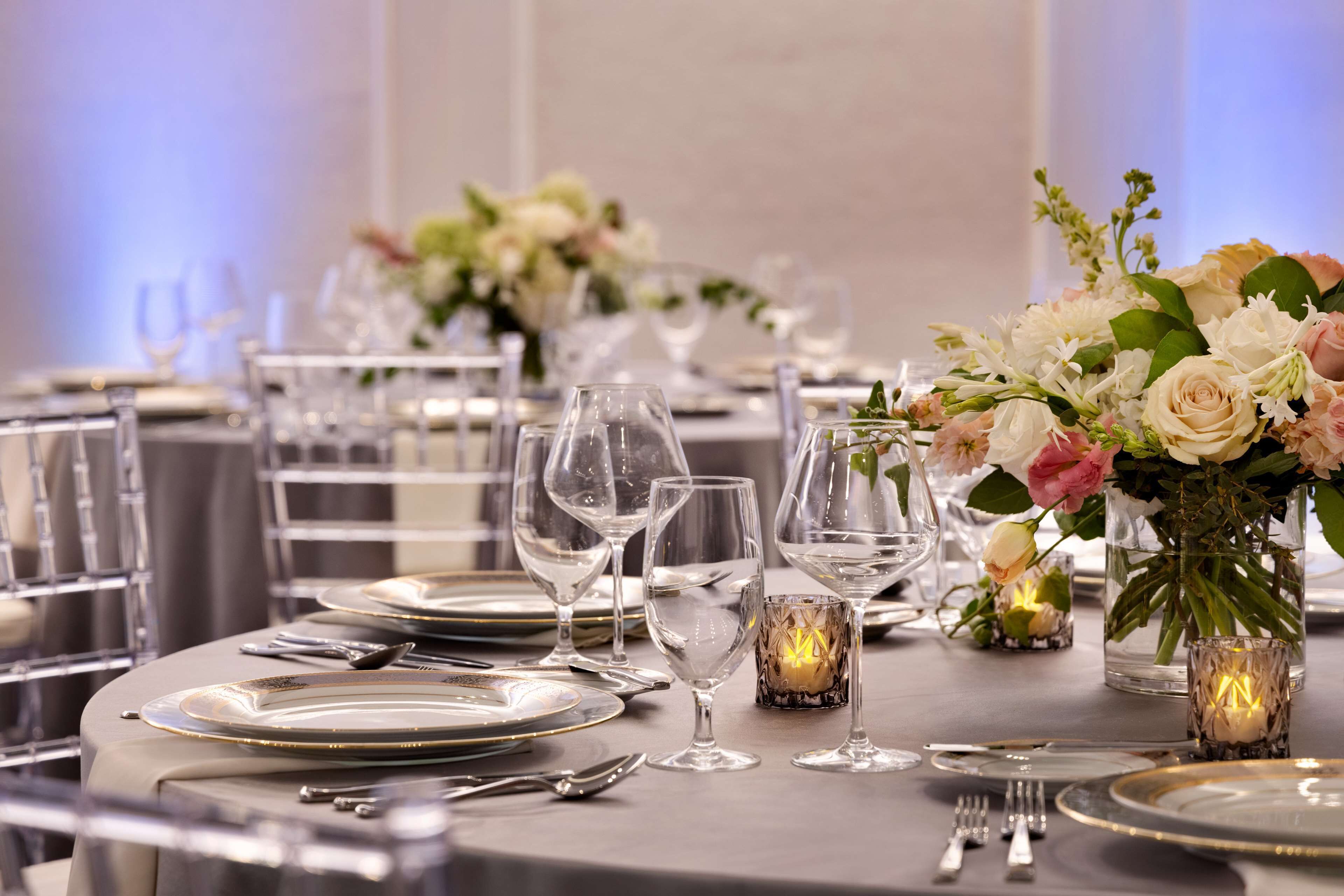 Una mesa dispuesta con cristalería transparente, platos y cubiertos, adornada con centros de mesa florales y velas encendidas.