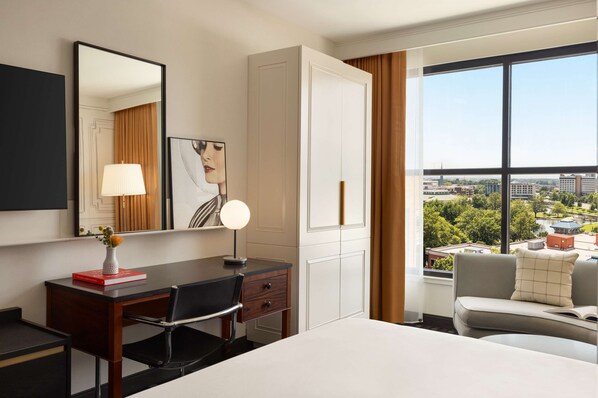 Habitación de hotel con una cama grande, un escritorio con silla, una lámpara de escritorio, un espejo y vistas de la ciudad a través de amplias ventanas.