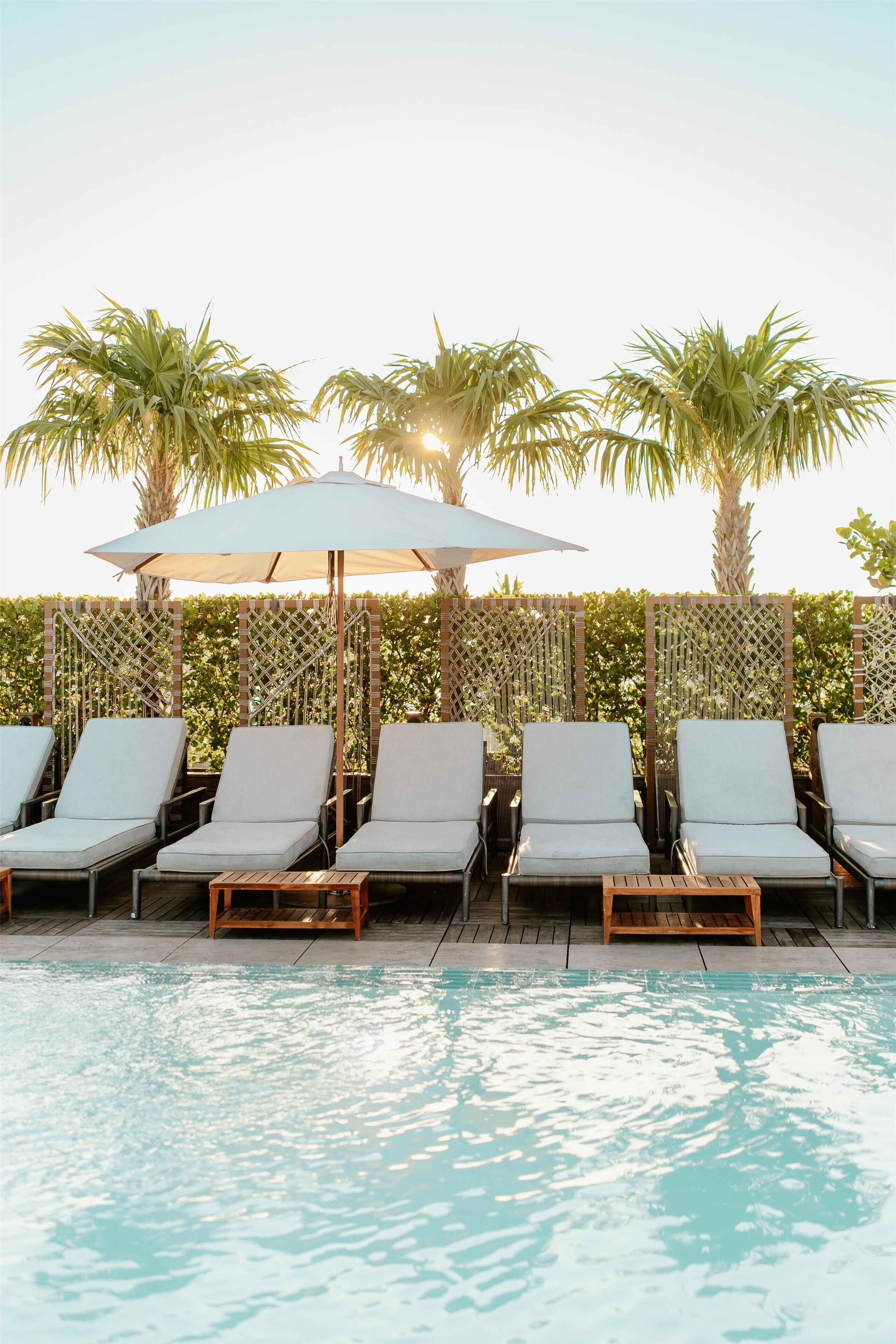 A poolside area with lounge chairs and umbrellas, surrounded by palm trees and a lattice fence.