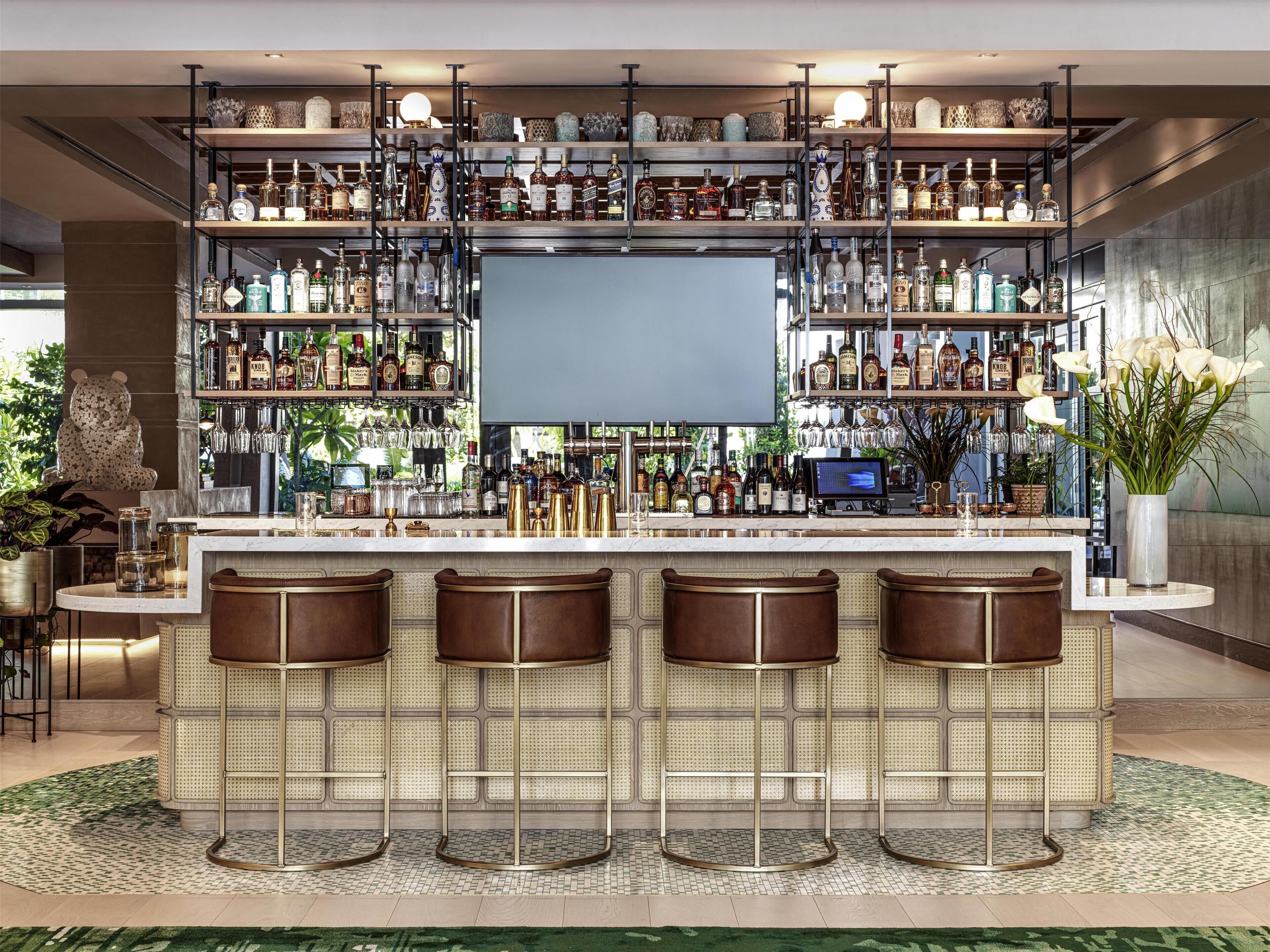 Zona de bar con varias estanterías llenas de botellas de licor, una pantalla grande y tres taburetes.