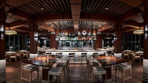 A bar area with wooden tables and chairs, a bar counter with shelves of bottles, and large windows providing a view of the city. - The Ray Hotel Delray Beach, Curio Collection by Hilton (Delray Beach)