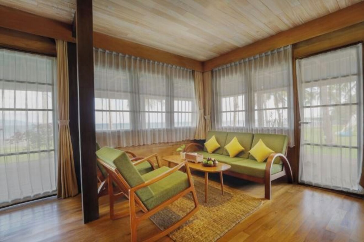 A room with a wooden ceiling, large windows with curtains, a green sofa with yellow cushions, and a wooden coffee table.