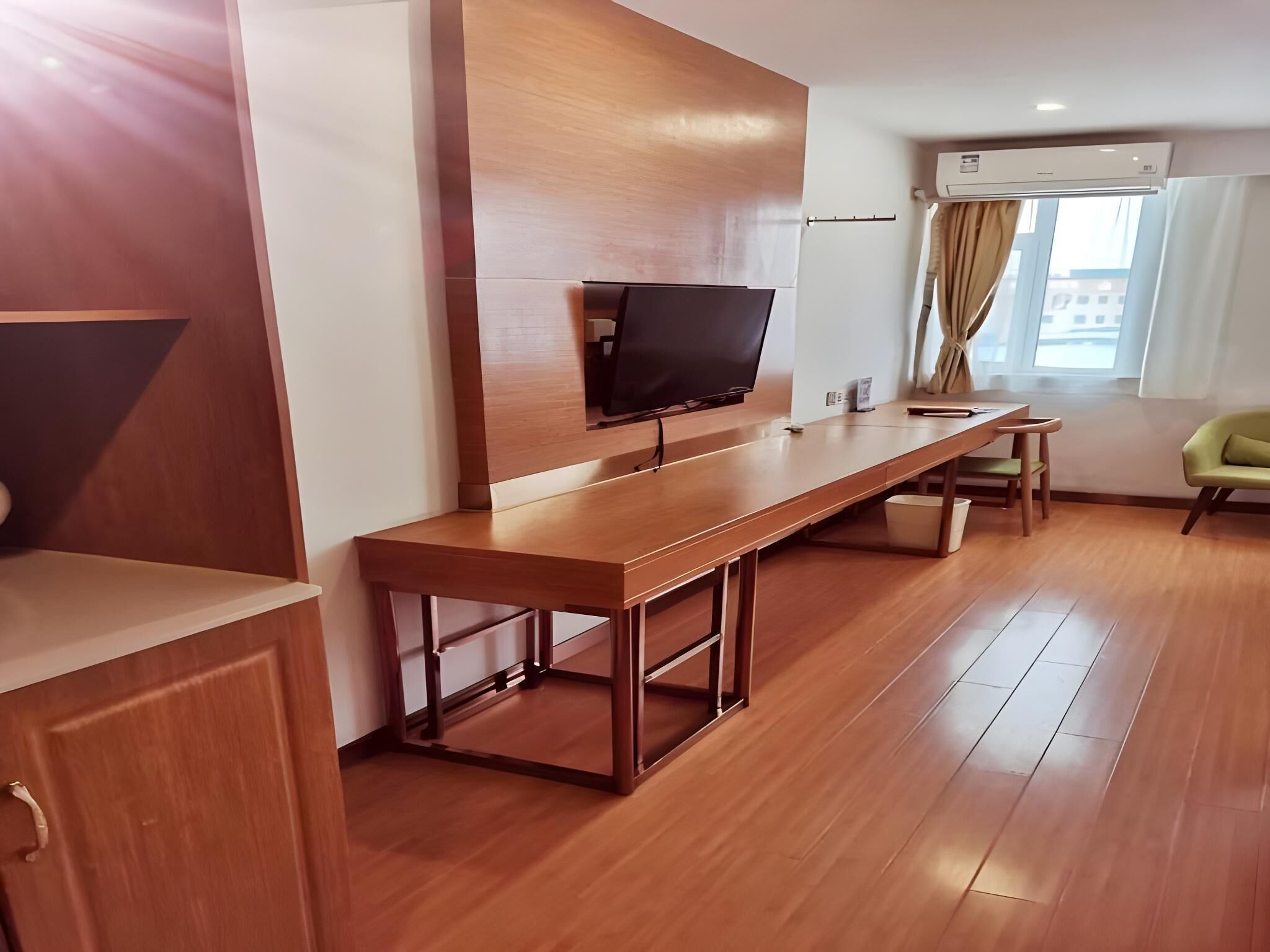 A modern hotel room with a wooden desk, a flat-screen TV, a window with curtains, and an air conditioning unit.