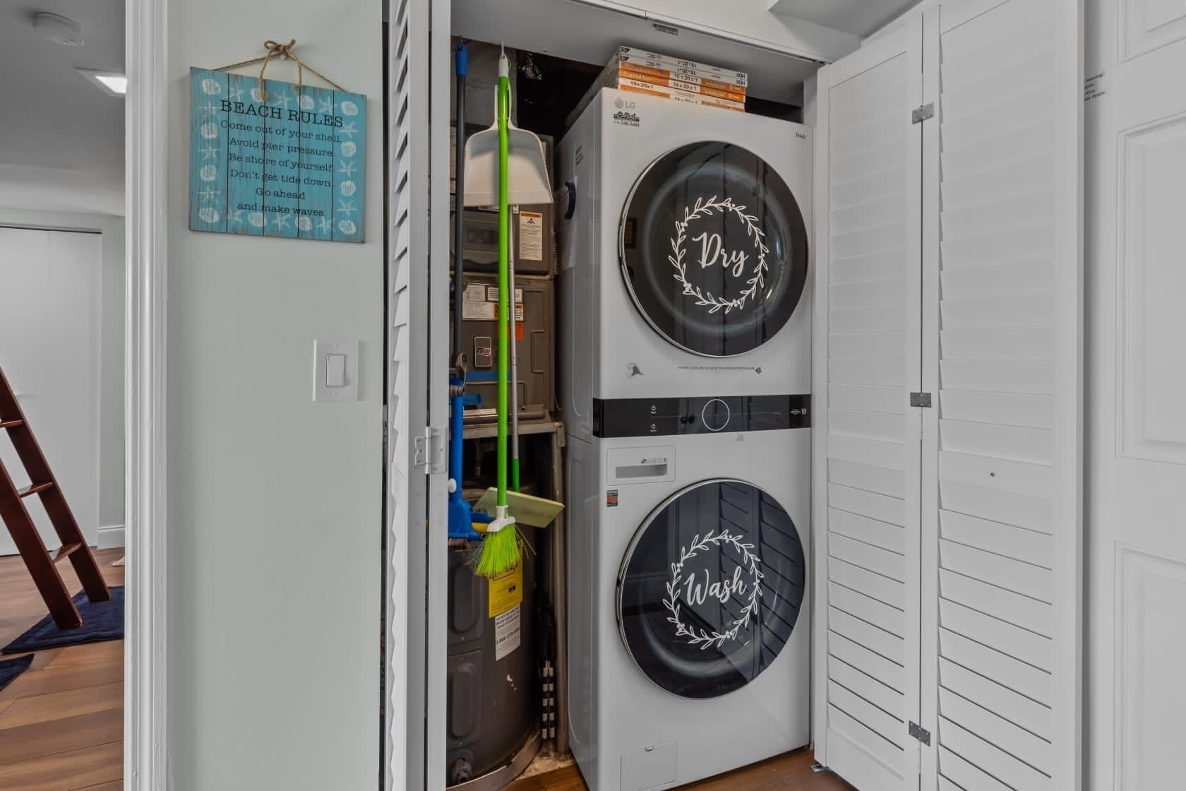 A laundry room with two stacked front-loading washing machines, a broom, and a sign with beach rules.