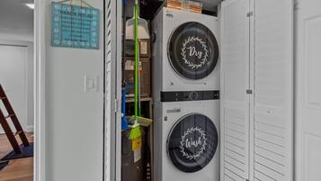 A laundry room with two stacked front-loading washing machines, a broom, and a sign with beach rules.