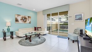 A modern living room with a light blue wall, a glass coffee table, a beige sofa, and a flat-screen TV. - EV_1445 – 3 Bed, 3 Bath with Private Pool and Access to Amazing Resort Amenities (clermont)