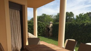 A covered patio area with a wooden table and chairs, a view of a garden with a stone structure, and a clear sky.