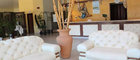 A lobby with two white leather sofas, a large terracotta vase with bamboo, and a reception desk with a clock.