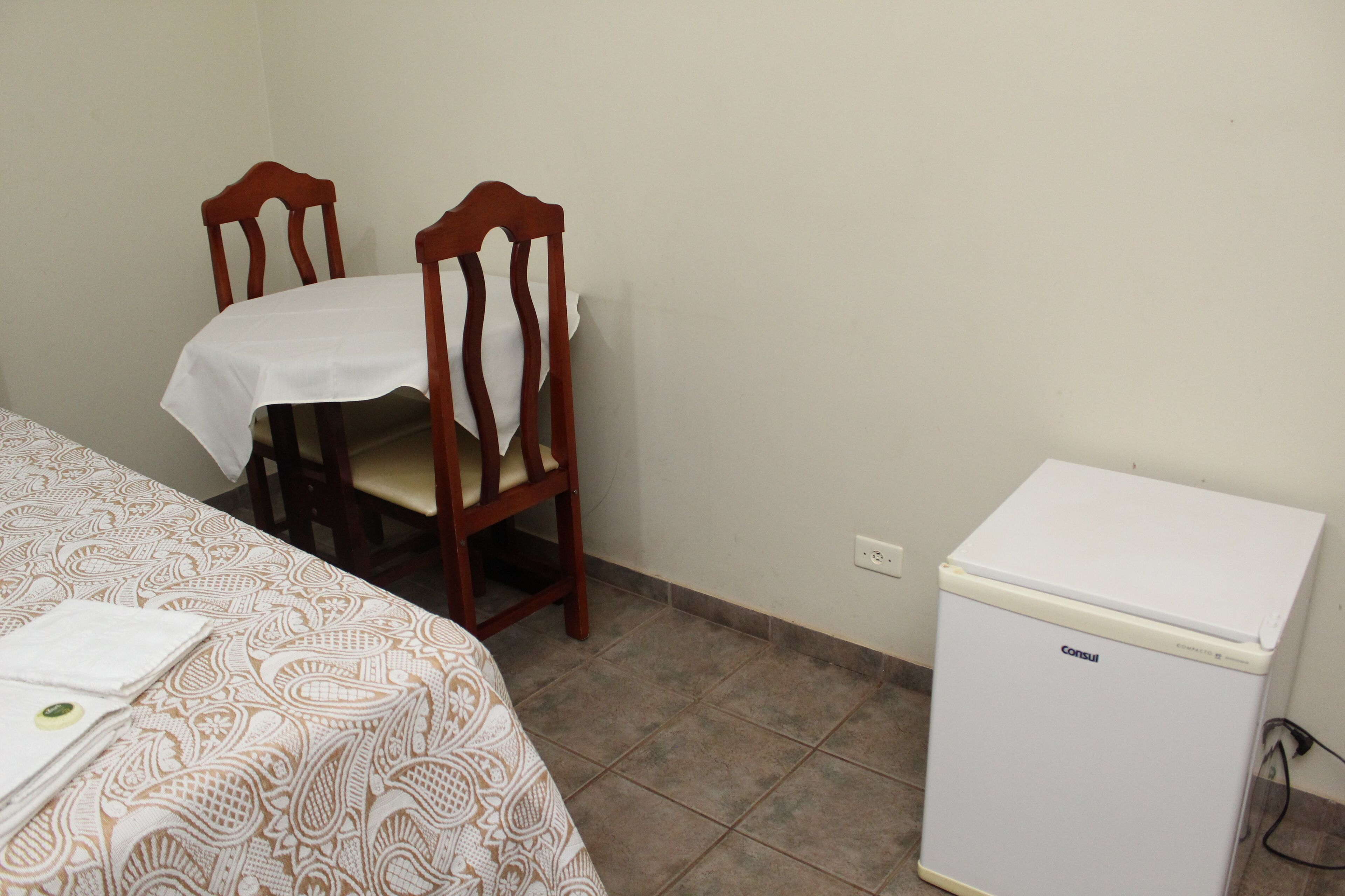 Une pièce comprenant un lit, une chaise en bois, un petit réfrigérateur blanc et une couvre-lit à motifs.