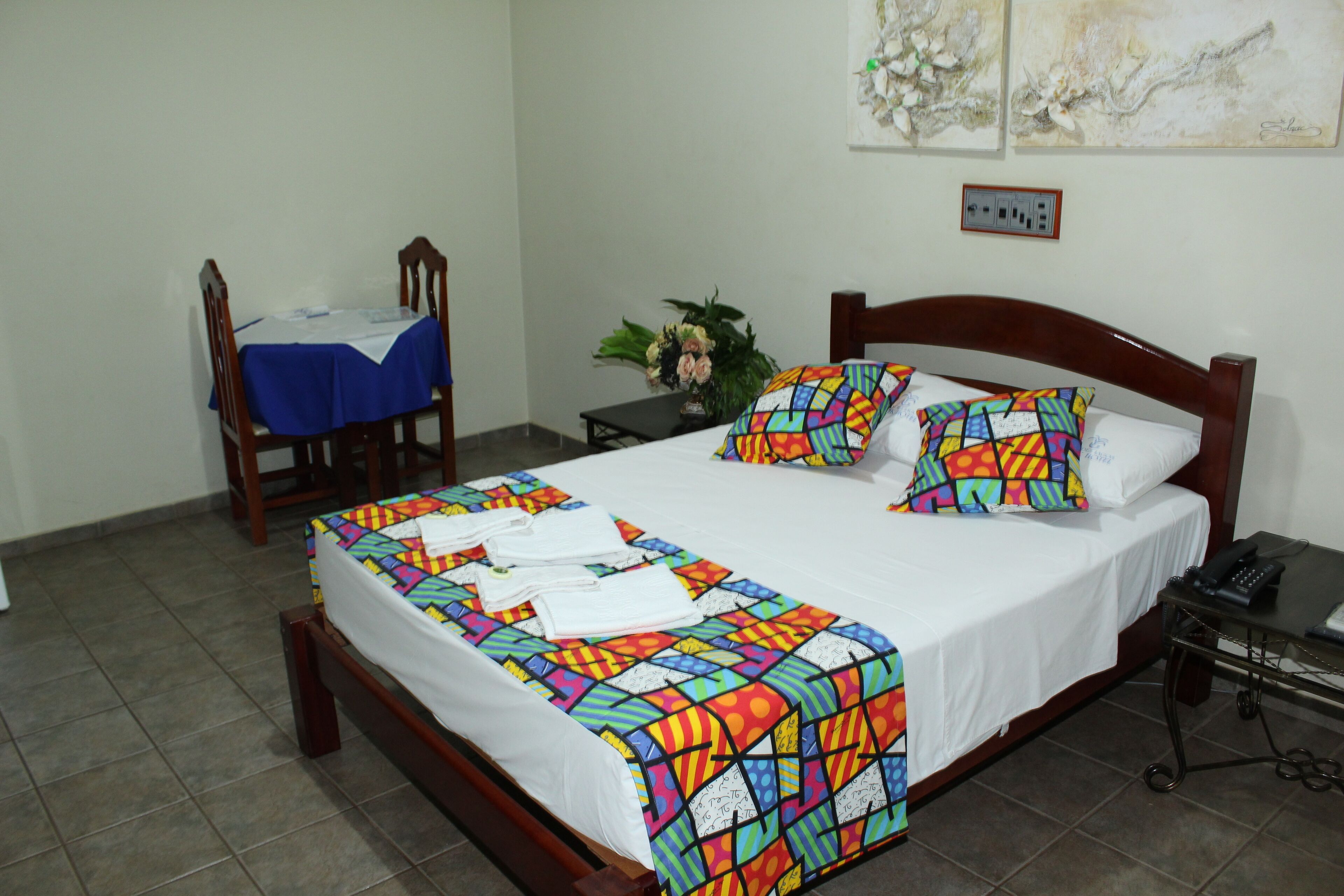 A neatly made bed with colorful bedding, a bedside table with a phone, and a chair with a table covered by a blue cloth.