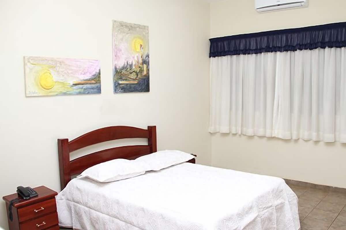 Un dormitorio con cama, mesita de noche, aire acondicionado de pared y dos cuadros.