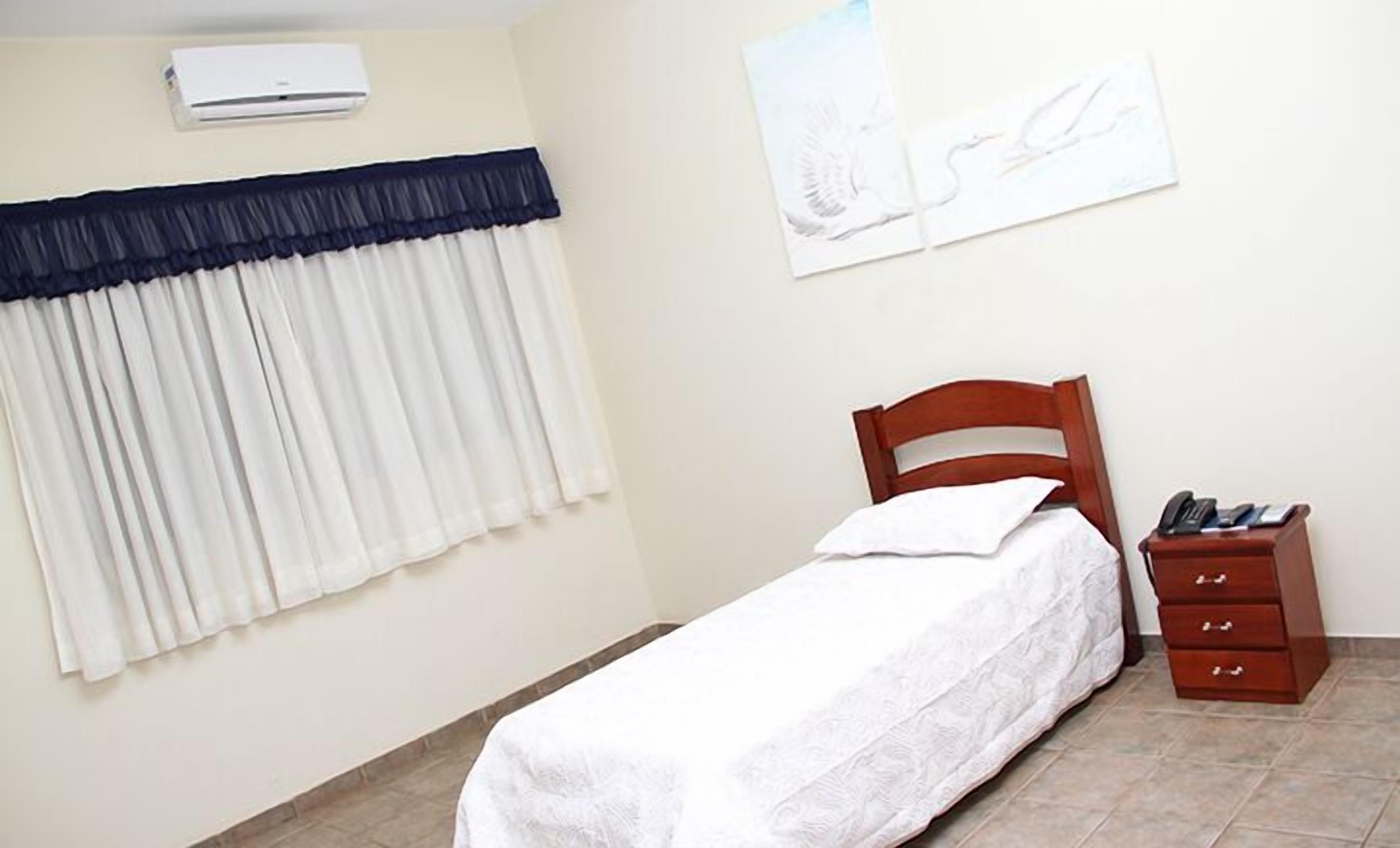 A bedroom with a single bed, a wooden nightstand with a phone, and a wall-mounted air conditioner.