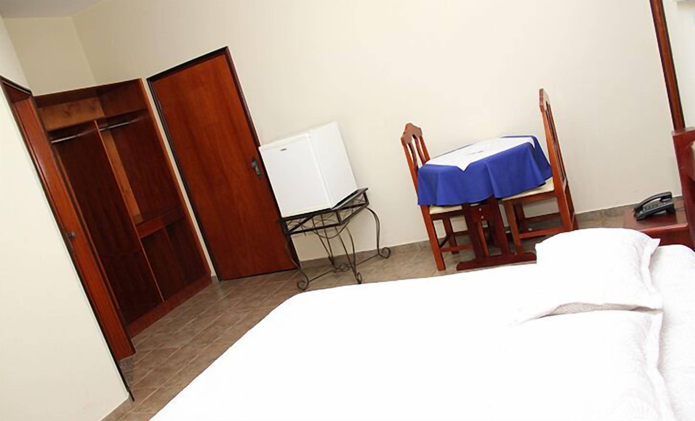 A hotel room with a bed, a desk with a blue tablecloth, a chair, a wardrobe, and a whiteboard.