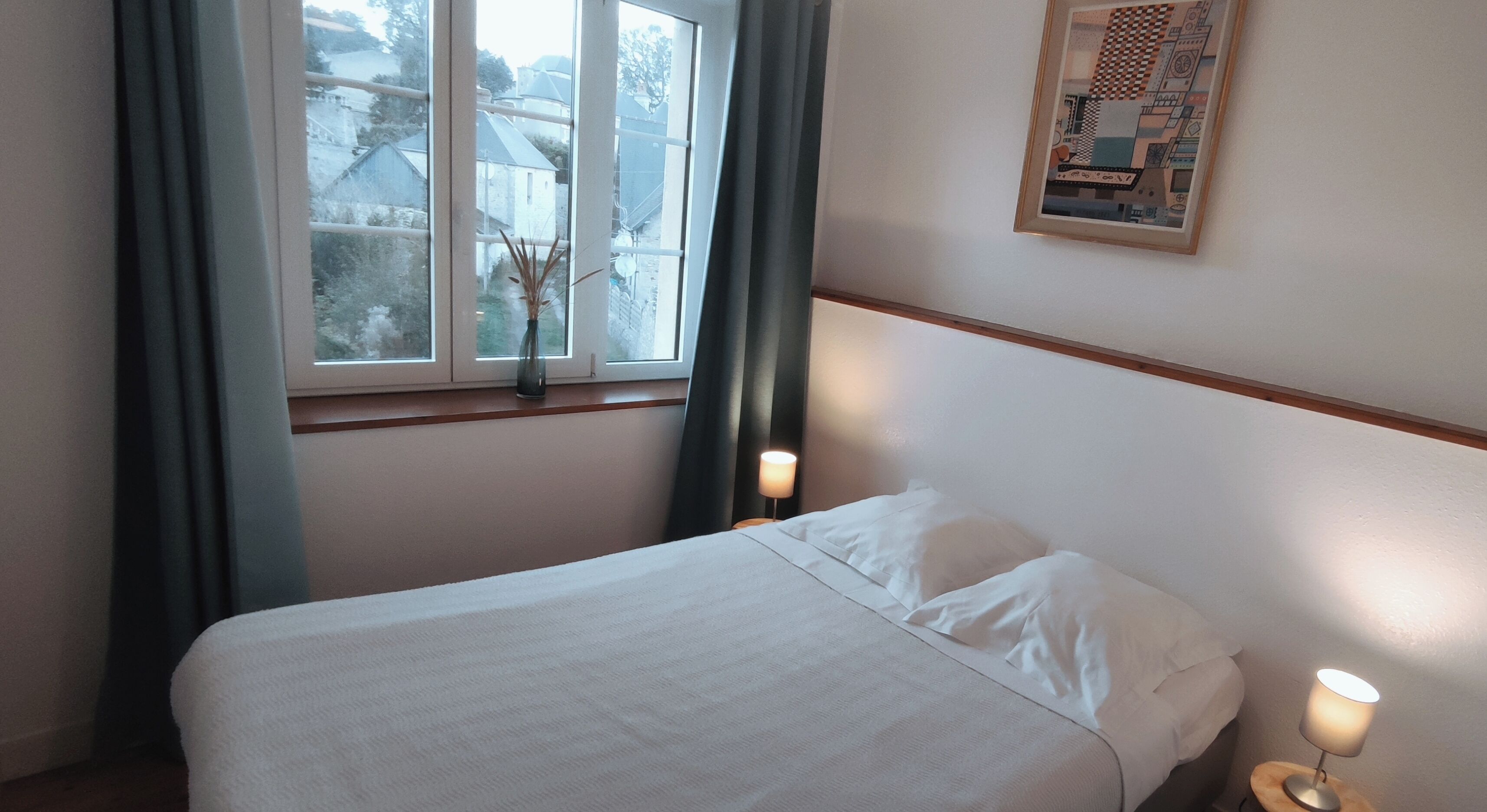 Ein ordentlich bezogenes Bett mit weißen Leinen in einem Hotelzimmer. Es gibt ein Fenster mit Vorhängen, ein gerahmtes Bild an der Wand und eine Nachttischlampe.