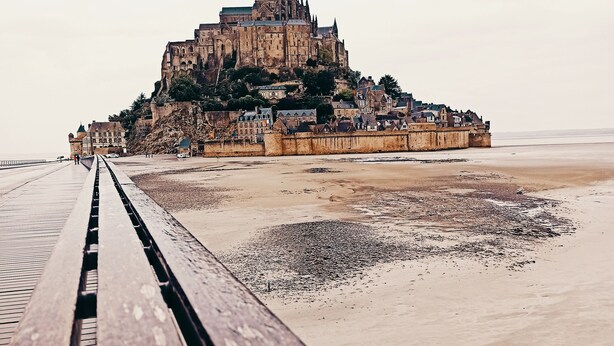 Un muelle de madera que conduce a un castillo histórico en un acantilado con vistas a una playa.
