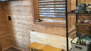 A wooden cabin interior with a bench, a window with blinds, a shelf with items, and a TV mounted on the wall.