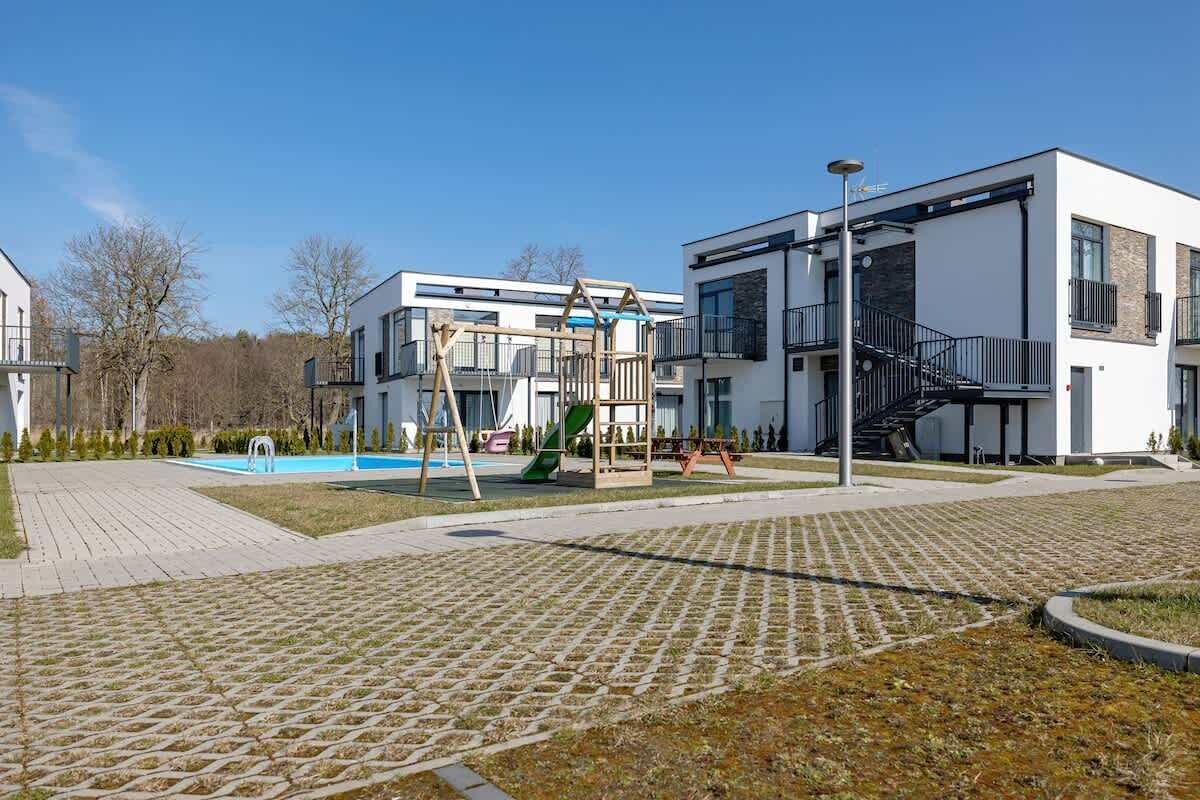 Ein Spielplatz mit Rutschen und Schaukeln, ein Schwimmbecken sowie moderne Gebäude mit Balkonen und einer Treppe.