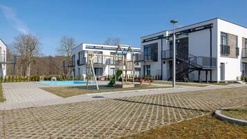 Ein Spielplatz mit Rutschen und Schaukeln, ein Schwimmbecken sowie moderne Gebäude mit Balkonen und einer Treppe.