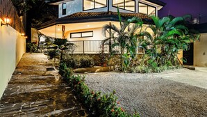 Una casa moderna de dos pisos con un camino de piedra que lleva a la entrada, rodeada de exuberante vegetación y un jardín bien cuidado.