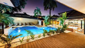 Una piscina con sillones de descanso, una terraza de madera y una cocina moderna visible a través de una puerta abierta.