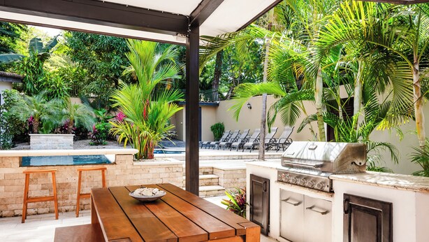Cocina al aire libre con una parrilla, taburetes de bar y una mesa de madera.