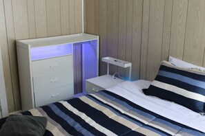 A bedroom with a bed, a bedside table, a nightstand, a wardrobe with a light, and a wall-mounted shelf.