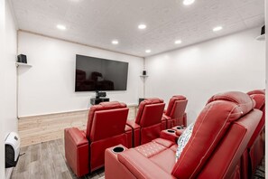A home theater room with red leather reclining chairs, a large flat-screen TV mounted on the wall, and a wooden floor.
