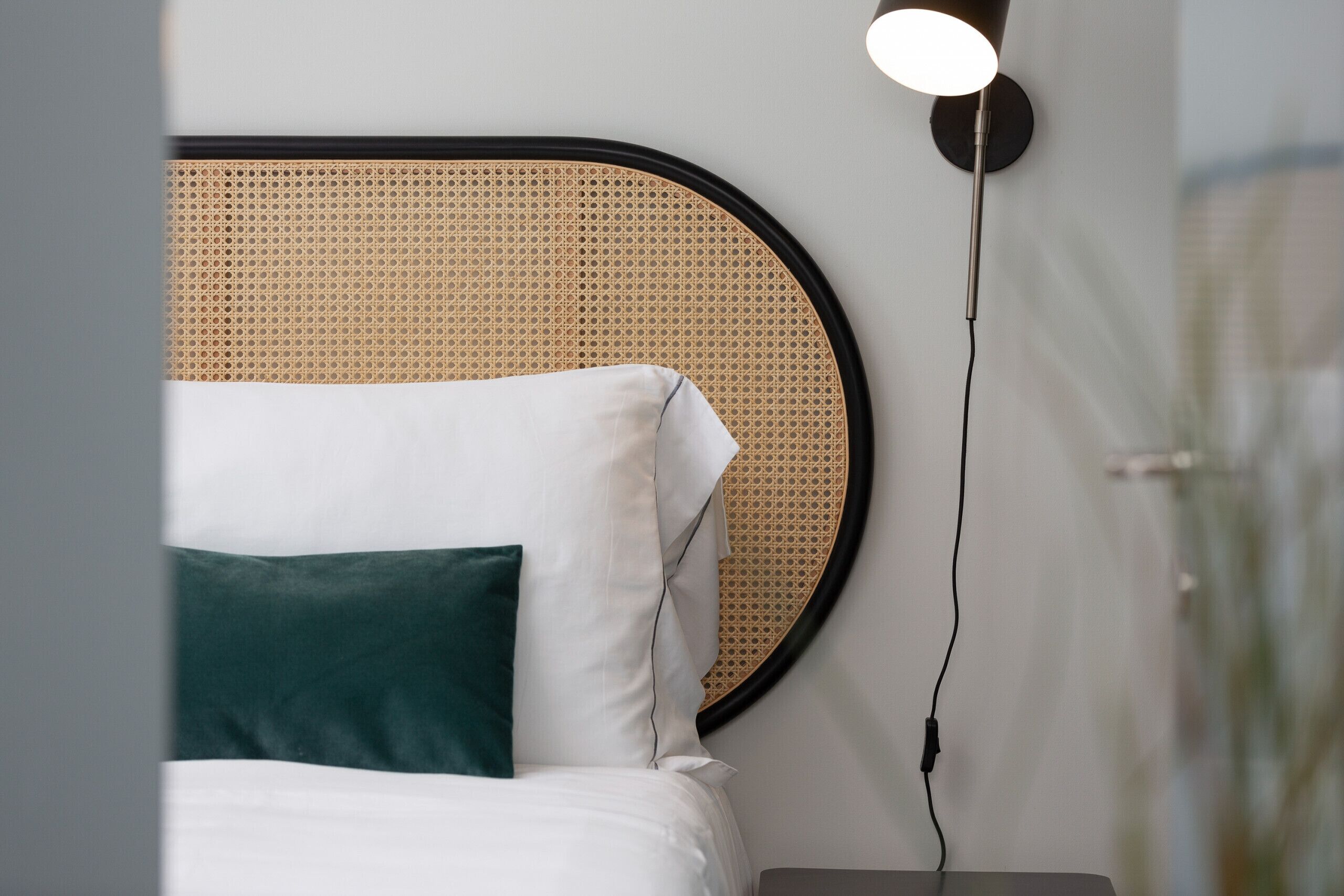 Una cama con almohadas blancas y verdes, un cabecero redondo tejido y una lámpara de noche.