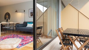 A modern living room with a glass partition, a dining area with wooden chairs and a table, and a sofa with a blue cushion.