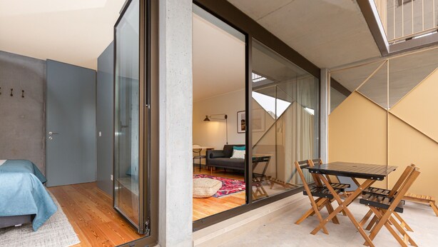 A modern interior with a glass door leading to a living area, a small dining table with chairs, and a bed with a blue cover.